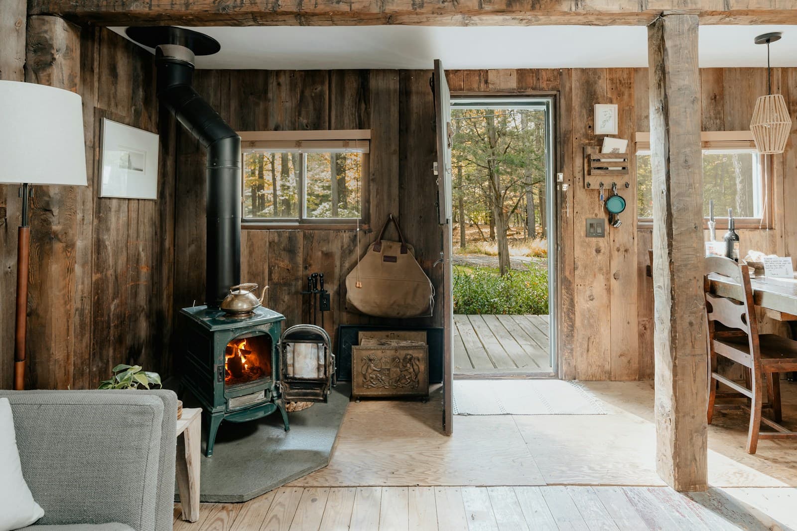 A green cast-iron wood stove with a kettle on top burns in a rustic wood-paneled cabin, with an open door onto autumn woods