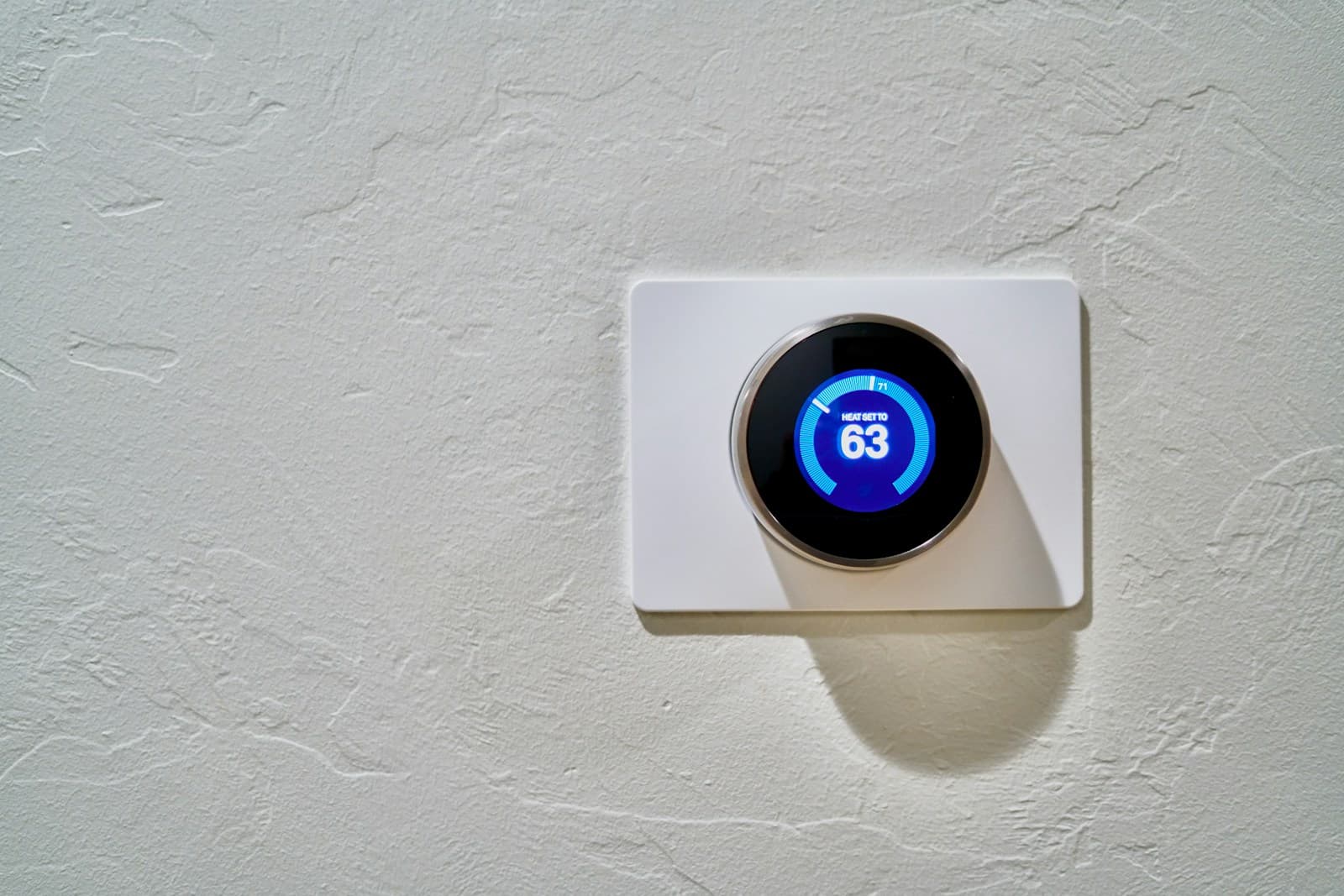 A round smart thermostat on a textured white wall, its blue display showing heat set to 63 degrees