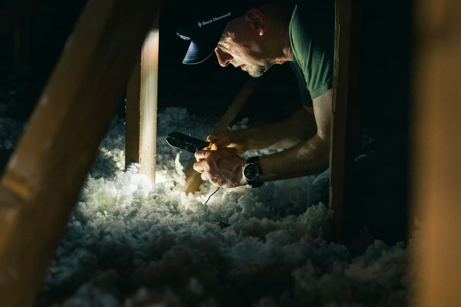 A man wearing a headlamp works among loose blown-in insulation between the rafters of a dark attic