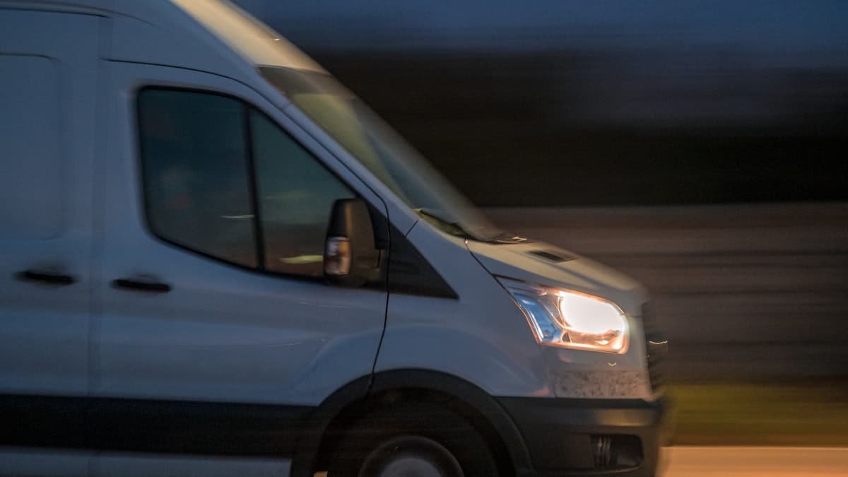 A white work van driving at dusk with its headlights on