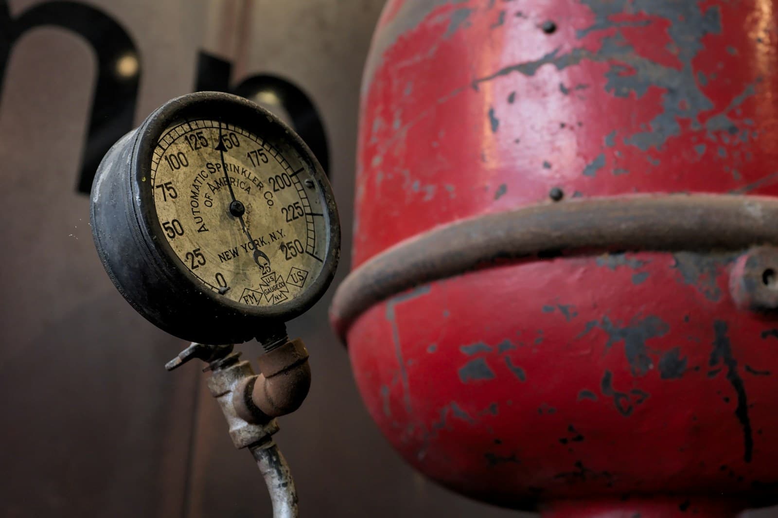 A vintage brass pressure gauge on a pipe beside a weathered red tank with chipped paint